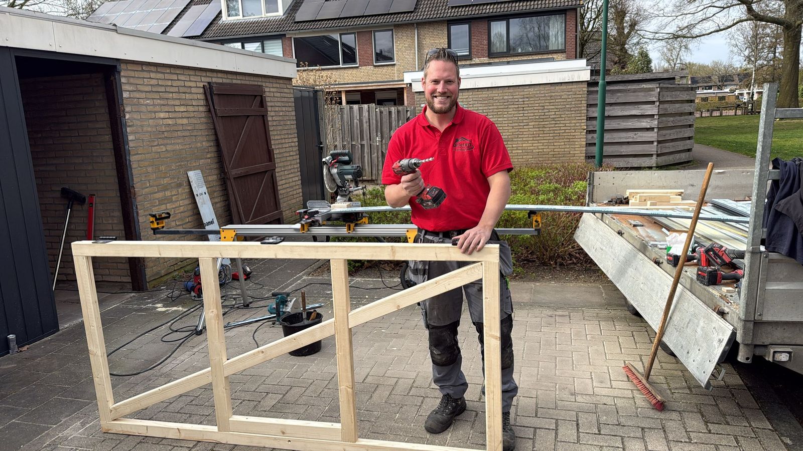 Vakman van Driesen Totaal Techniek in rood werkshirt met boormachine, achter een net getimmerd houten frame op een oprit