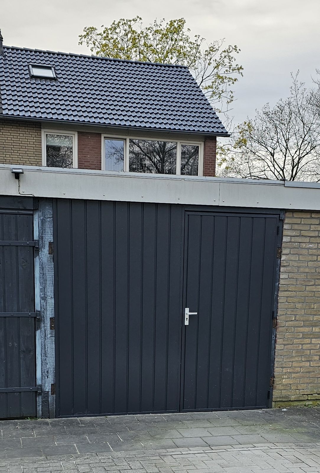 Nieuwe antraciet garagedeuren met een loopdeur, naast een oude houten deur
